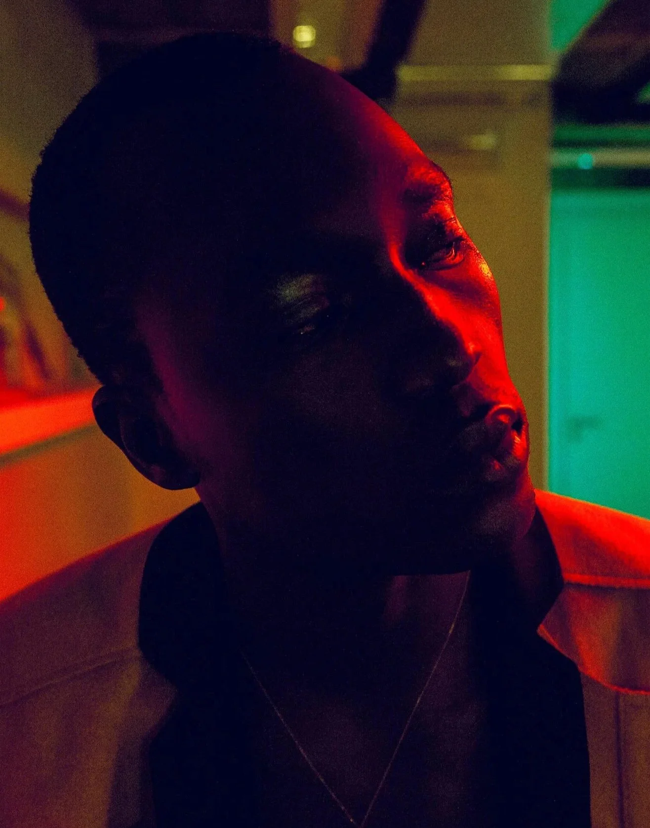A person with short hair in a dimly lit room with multicolored lighting, including red and green hues.