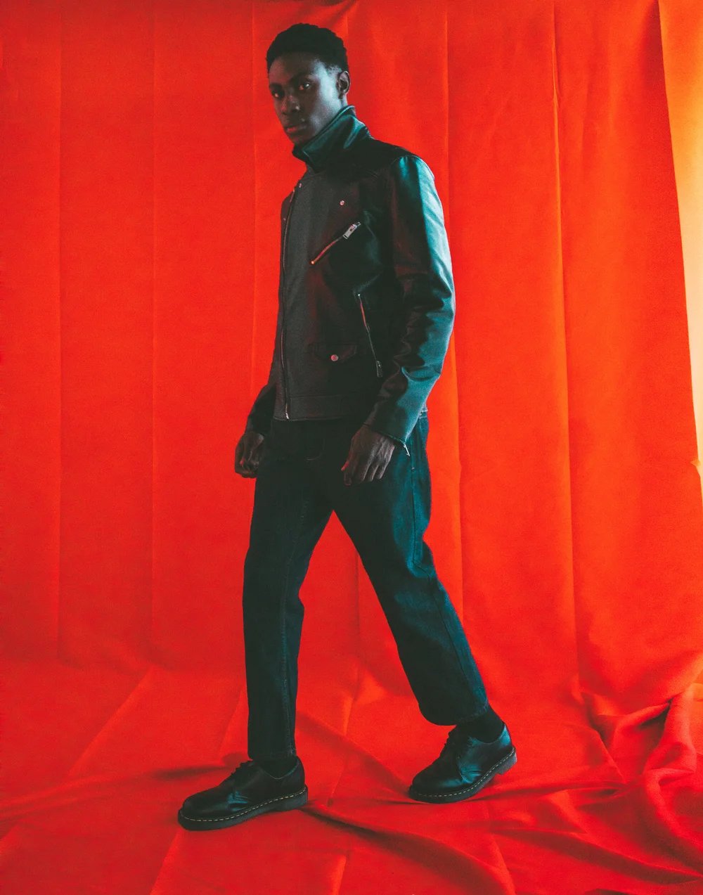 A young man with dark skin and short curly hair, wearing a black leather jacket, black pants, and black shoes, standing against a vibrant red curtain background.