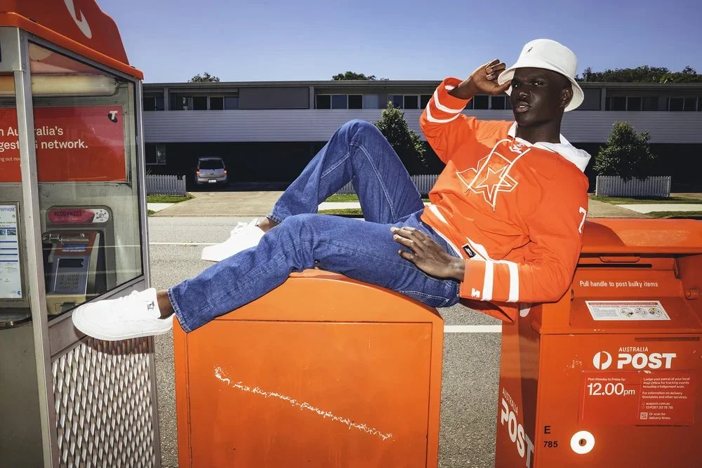 A person in a red and white hoodie, white hat, blue jeans, and white sneakers lounging on an orange Australia Post mailbox while talking on a phone in an outdoor parking lot.