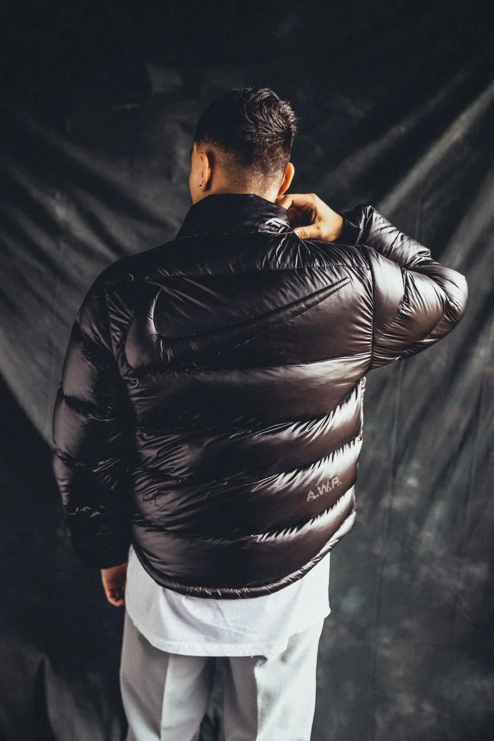 A person wearing a black, shiny, puffy jacket with a Nike logo on the back, standing against a dark, textured background, viewed from behind.