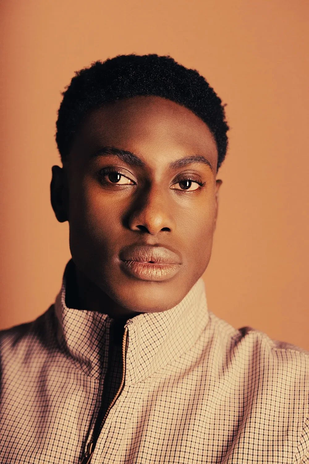 Close-up portrait of a young person with short curly black hair, wearing a beige checkered high-collared jacket, against a warm beige background.