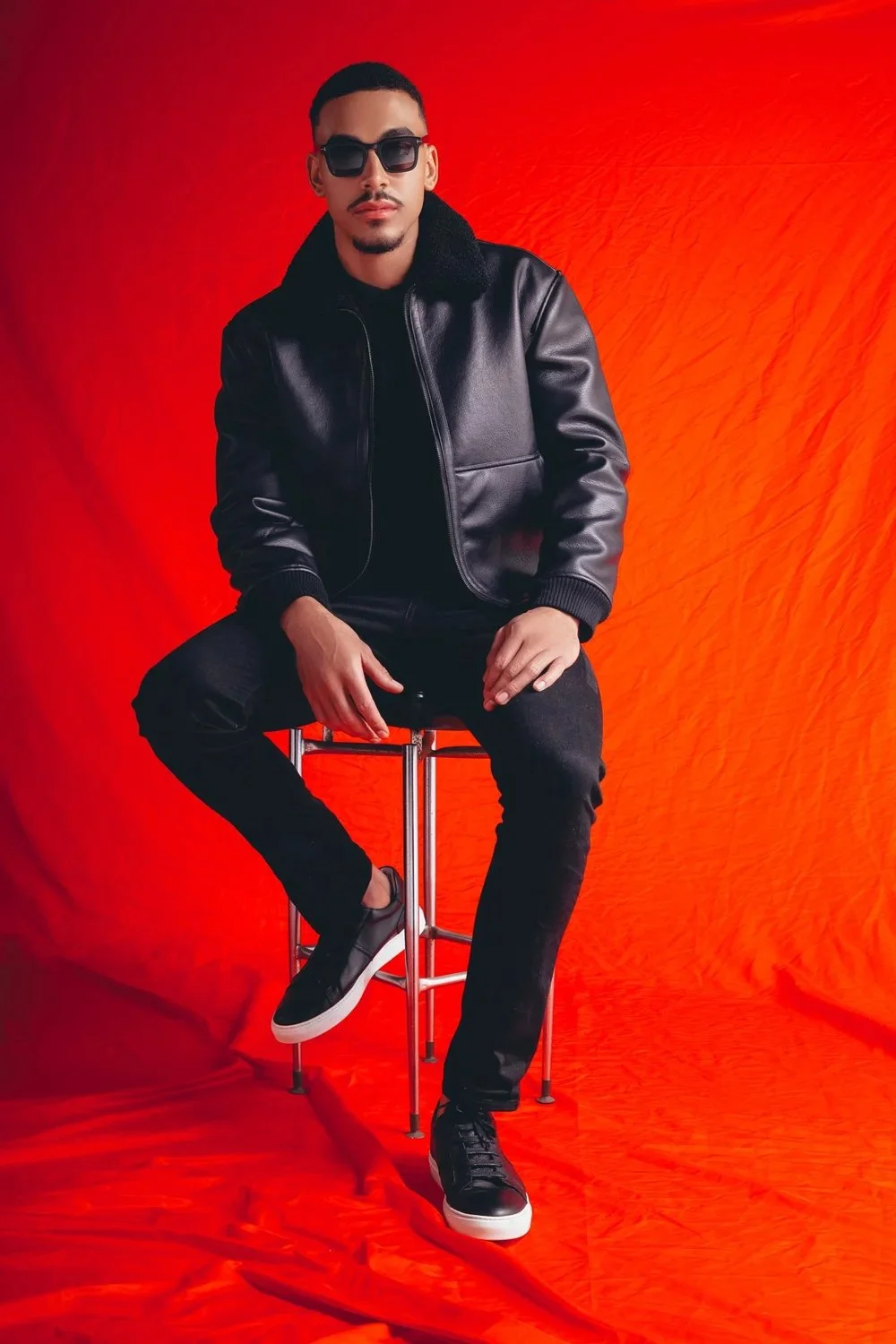 A man sitting on a stool in front of a bright red backdrop, wearing sunglasses, a black leather jacket with a fur collar, black pants, and sneakers.