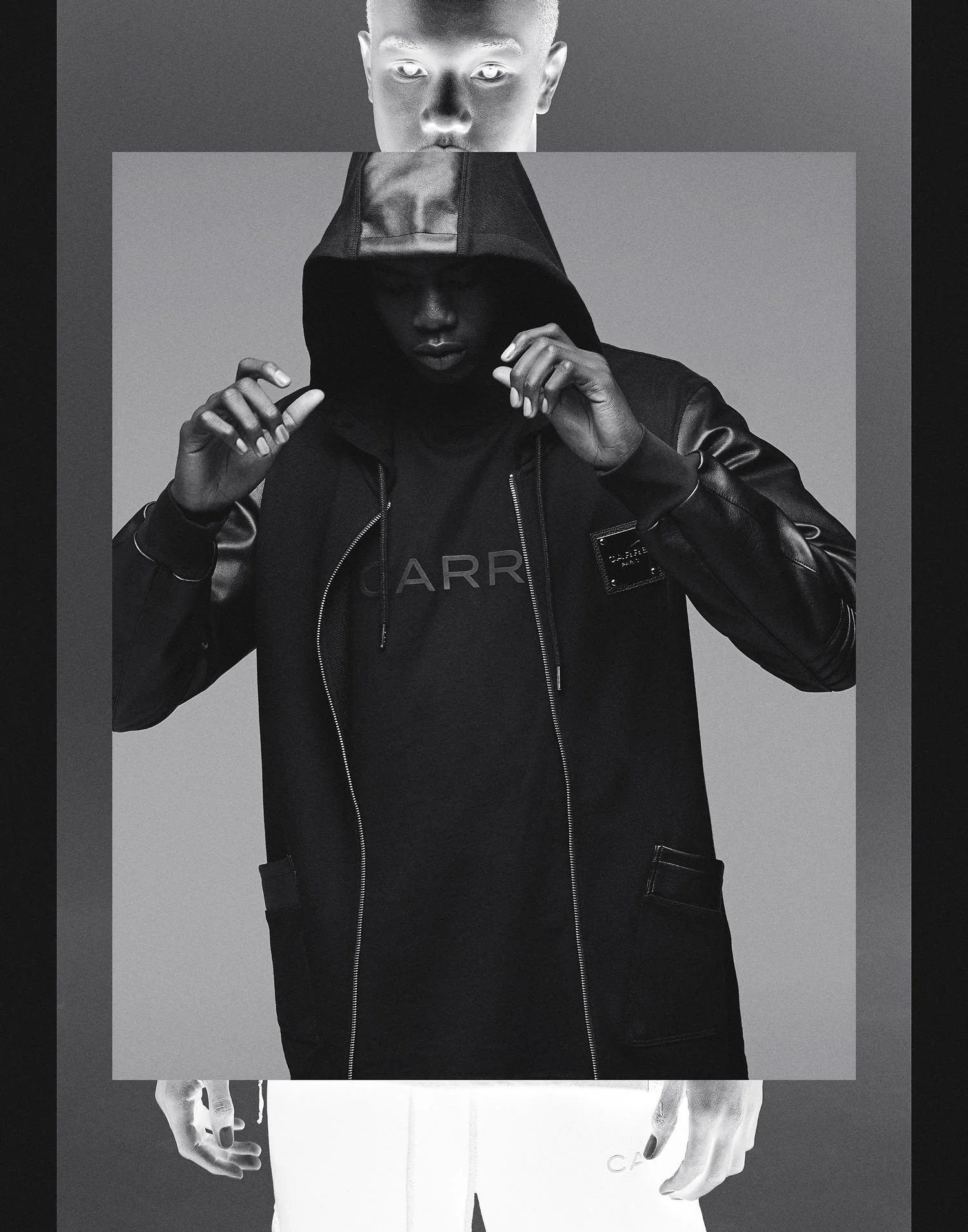 Black and white fashion photo of a person wearing a hooded jacket and a T-shirt, with their hands adjusting the hoodie, against a gray background.