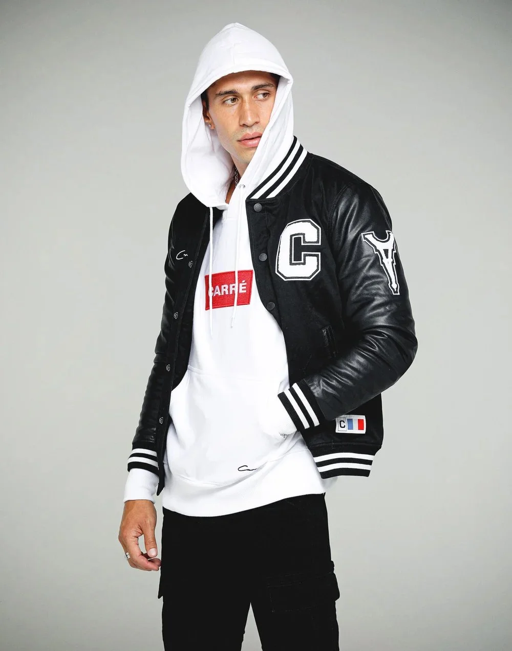 A young man wearing a white hoodie with a red logo, layered under a black leather jacket with patches, and black pants, standing against a plain background.