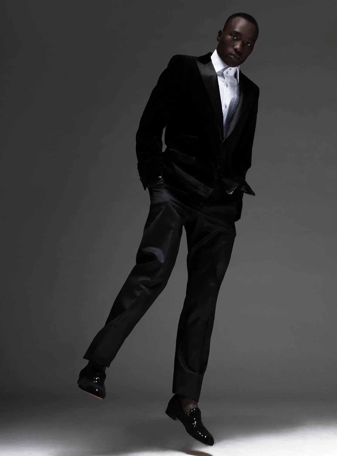 A man wearing a black tuxedo with a white shirt and black shiny shoes, posing against a plain dark background.