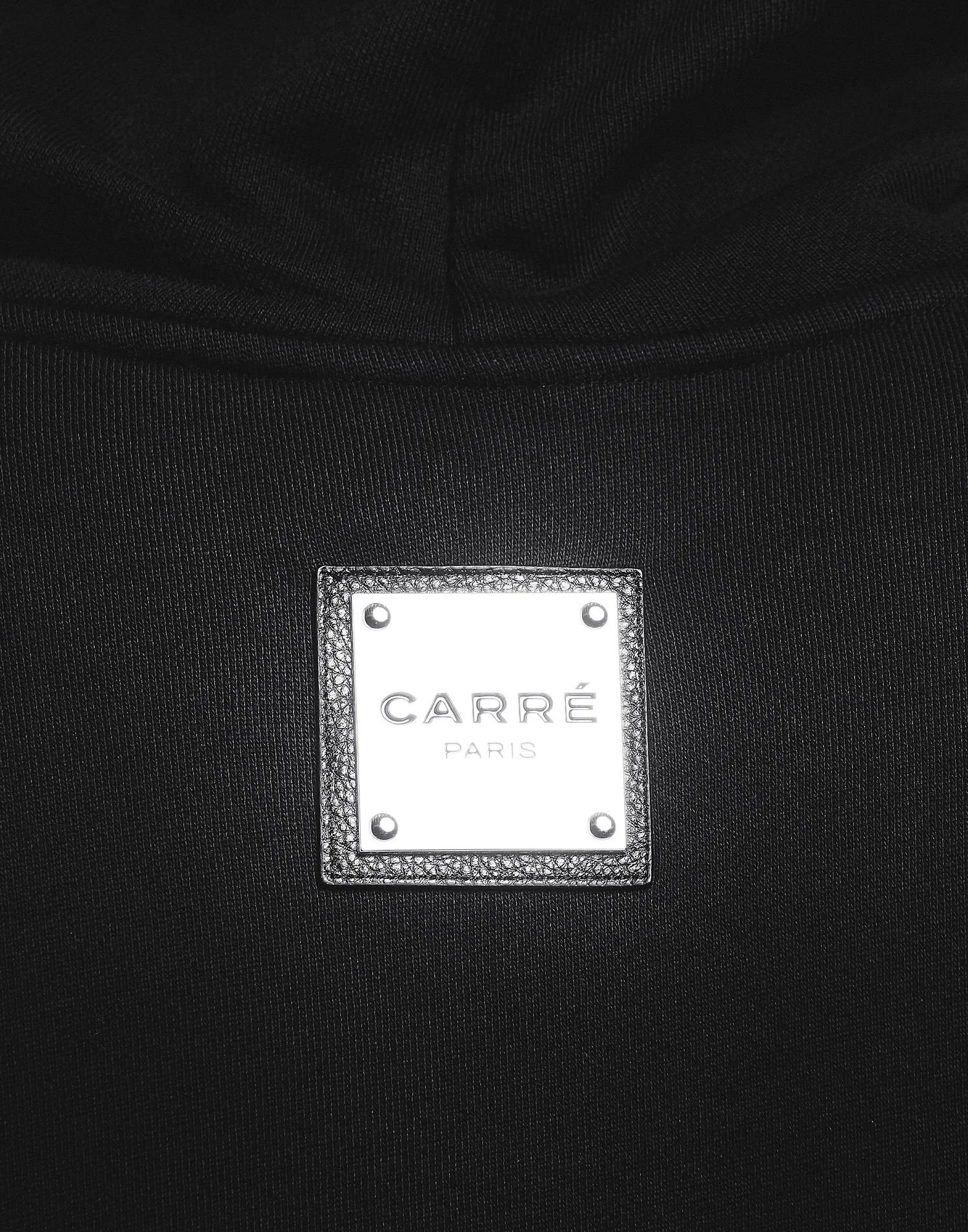 Close-up of a black garment with a silver square label reading 'CARRÉ PARIS' attached to the fabric.