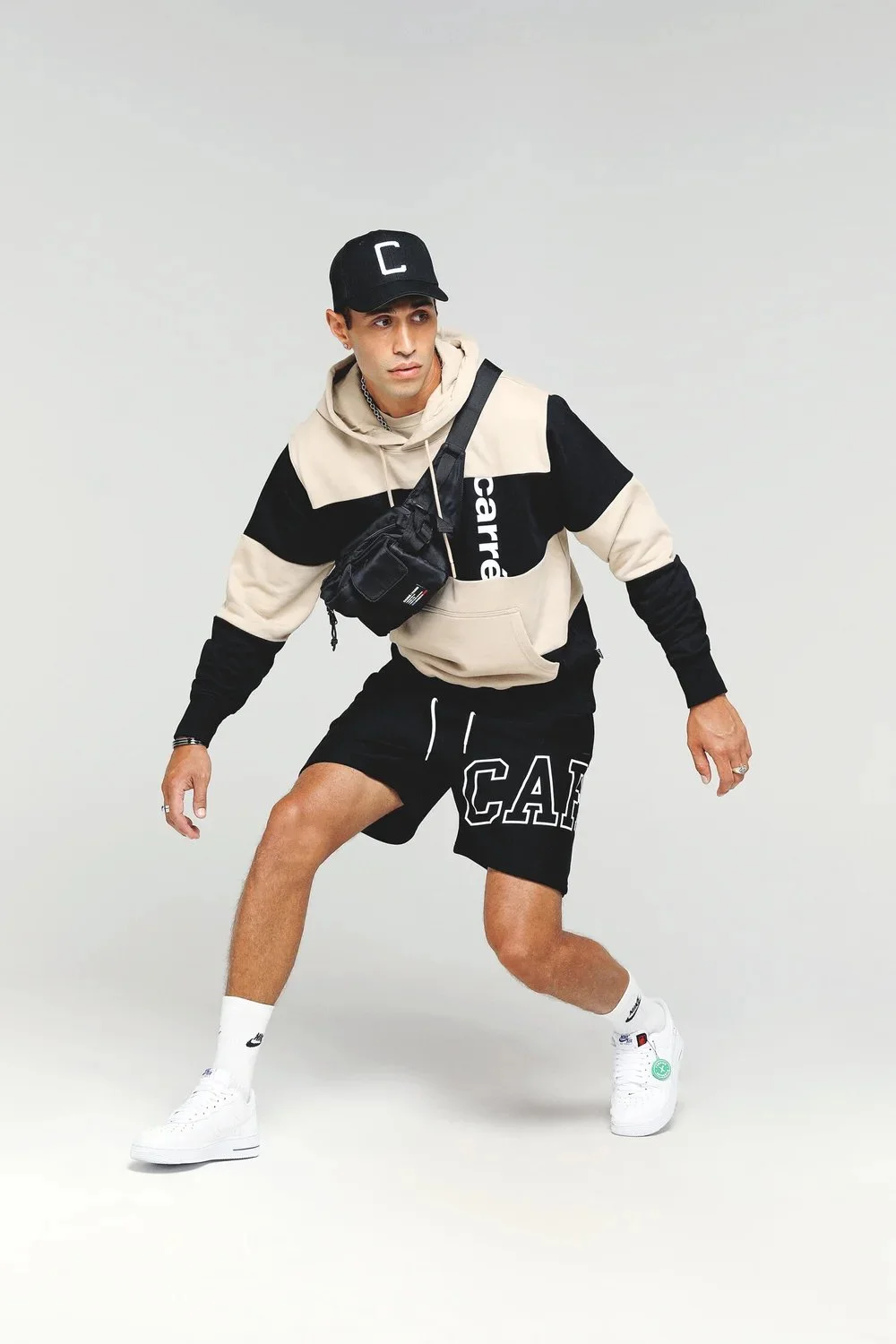 A young man in a beige and black hoodie, black shorts, white Nike sneakers, and a black cap is crouching with one foot forward in a studio with a plain light background.