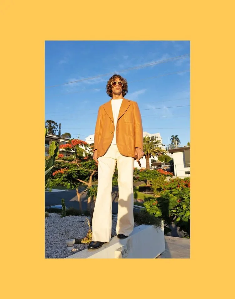 Person standing outdoors on a small pedestal, wearing a tan blazer, white shirt, cream wide-leg pants, black shoes, sunglasses, with a sunny blue sky and residential houses with palm trees in the background.