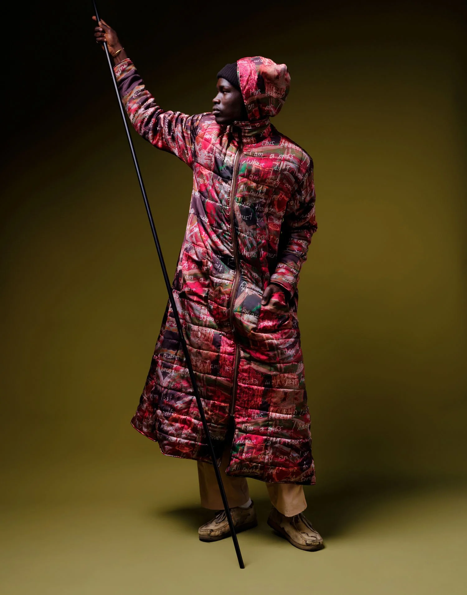 A person dressed in a long, quilted red patterned coat with writing on it, wearing beige pants, hiking boots, and a head covering, holding a walking stick against a dark background.