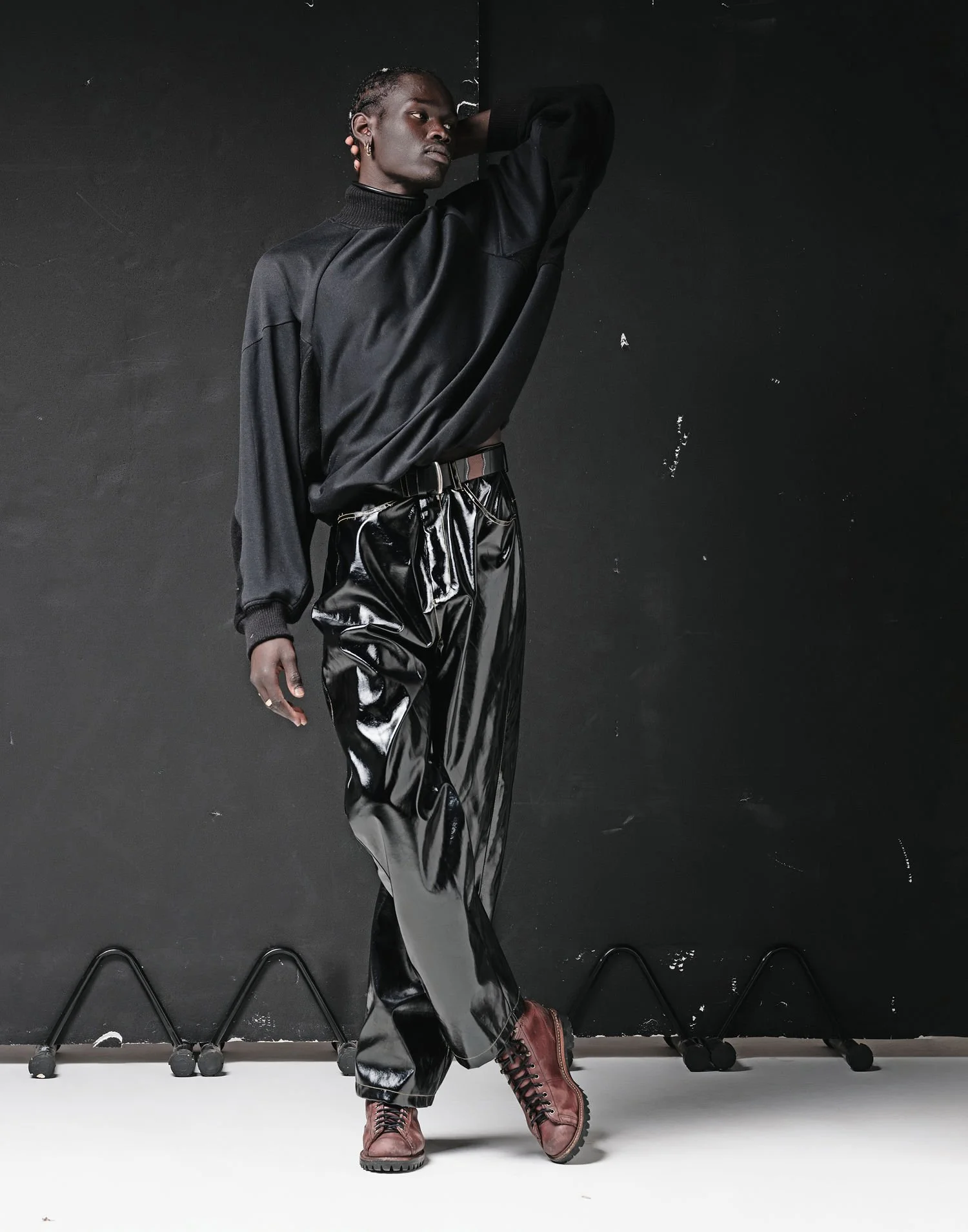 A person with dark skin and braided hair, wearing a loose black sweater, shiny black pants, and brown boots, standing against a black background in a studio.