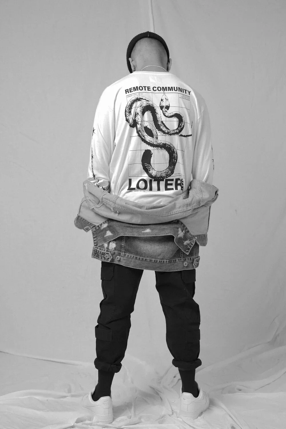 Back view of a person wearing streetwear, including a graphic jacket with a snake illustration and text about remote community and looters, black pants, white sneakers, and a beanie, standing against a plain wall.