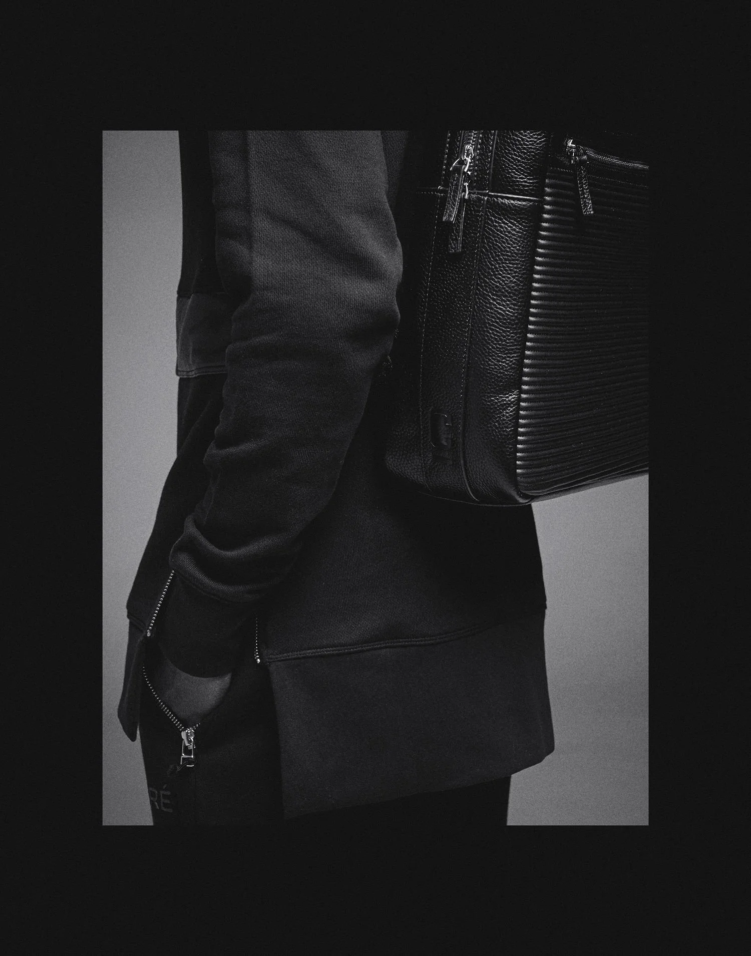 Close-up of a person wearing black clothing, holding a textured black bag with a zipper, and wearing a zippered jacket or top.