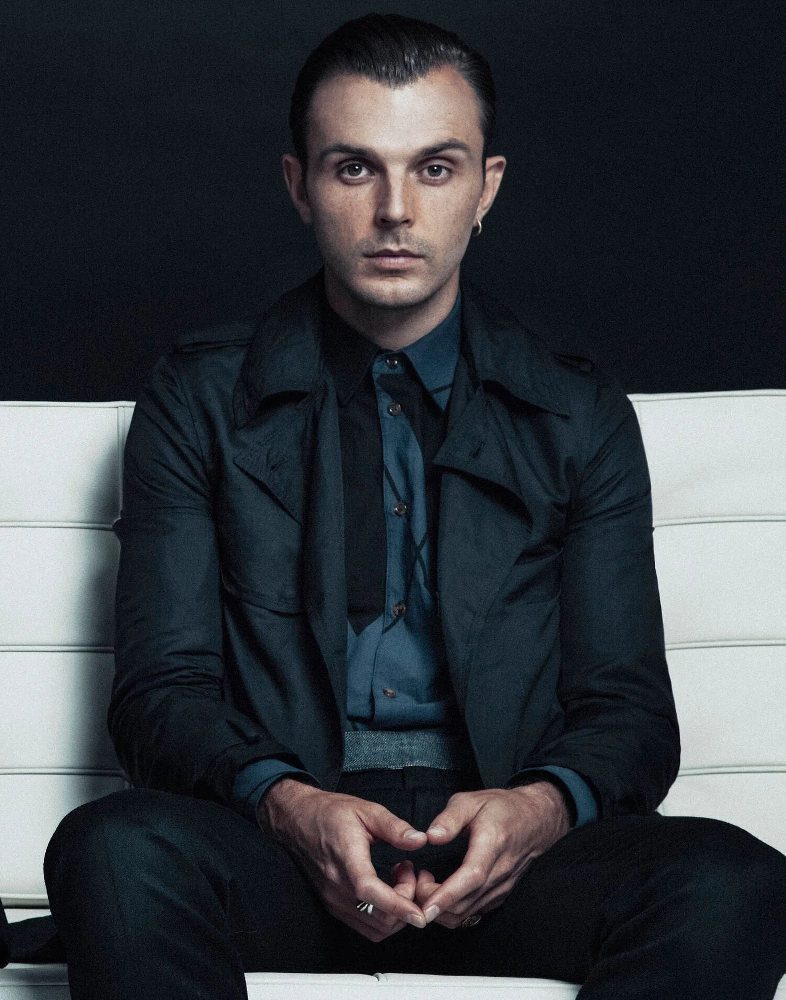 A young man with dark hair, wearing a dark jacket and blue shirt, sitting on a white bench against a black background, looking directly at the camera, with his hands clasped.
