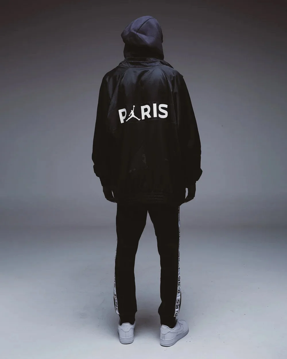 Person standing with their back to the camera, wearing a black hooded jacket with 'PARIS' and a Jumpman logo on the back, black pants with 'PARIS' on the side, white sneakers, in a simple studio setting with a gradient background.