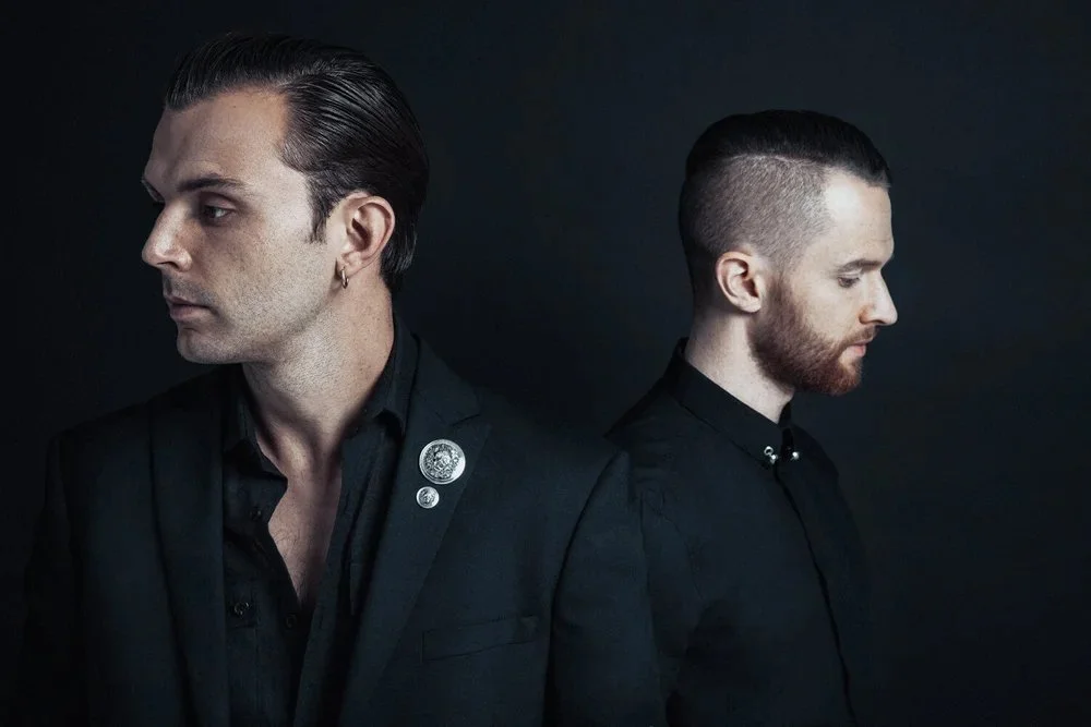 Two men with slicked-back hair and facial hair, dressed in black, standing back to back against a dark background, looking away from each other.
