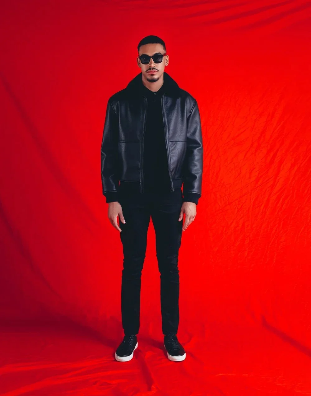 A man wearing sunglasses, a black leather jacket, black pants, and black sneakers with white soles, standing against a red backdrop.