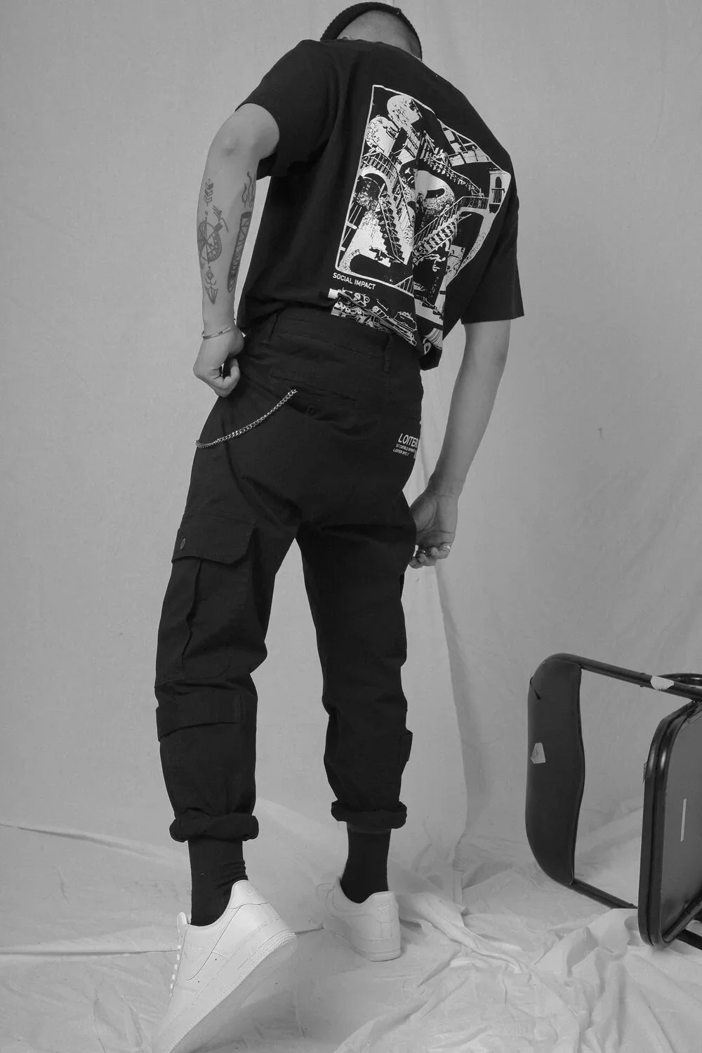 Person wearing a graphic black t-shirt, black pants with chain, white sneakers, standing on a white sheet, with camera equipment on the floor.