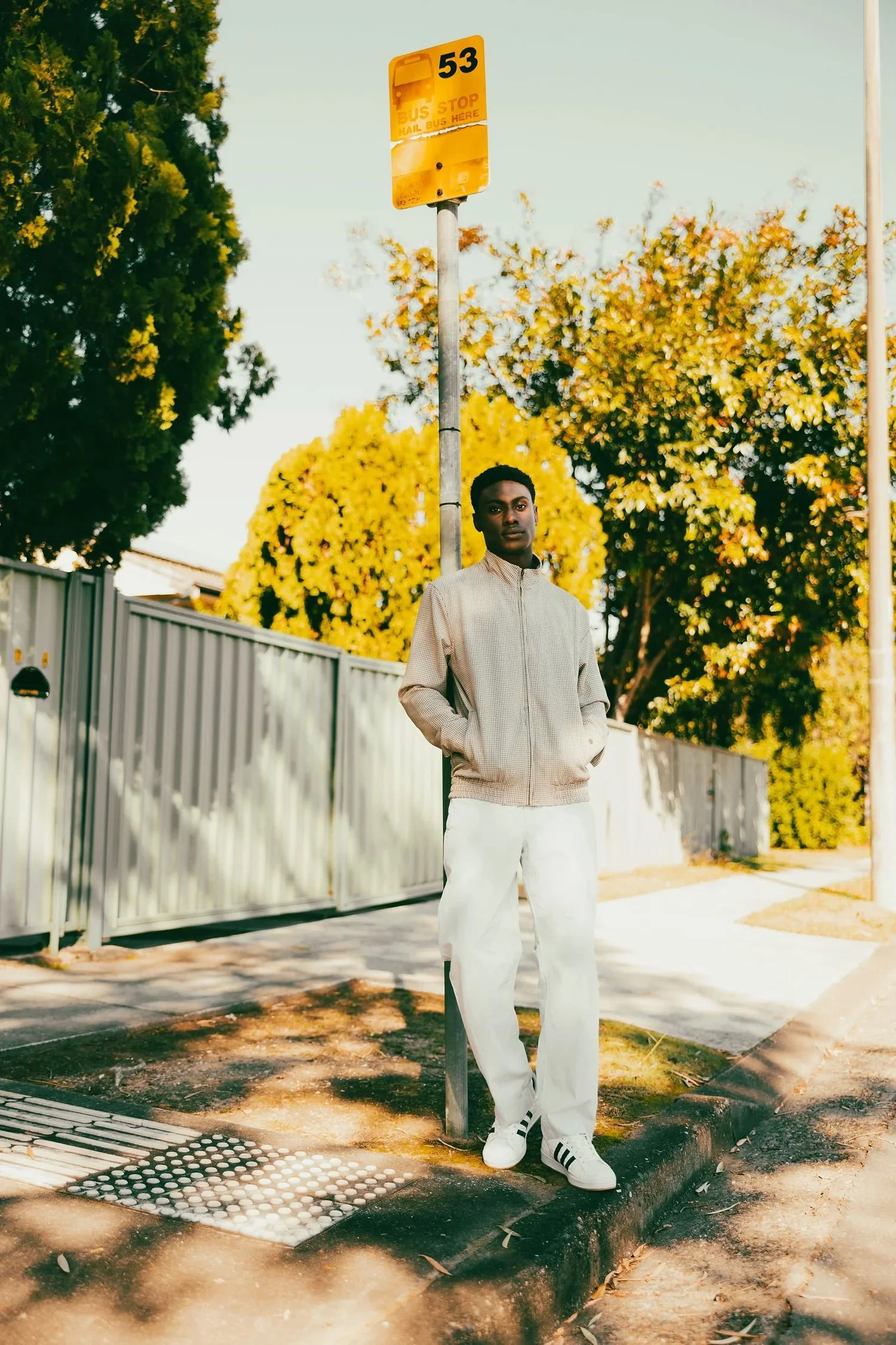 Young man standing beside a bus stop sign on a sidewalk with a background of trees and a fence.