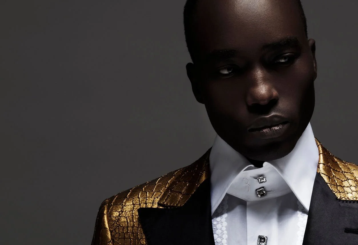 A dark-skinned person is wearing a black suit with gold textured epaulets on the shoulders, a white shirt with decorative buttons, and has a serious expression with a gray background.