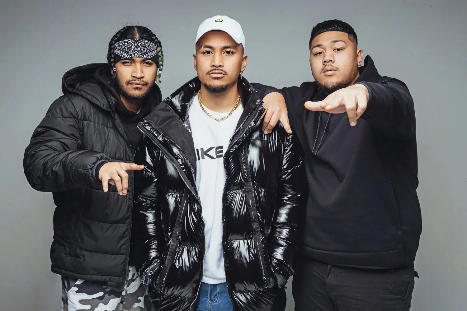 Three young men posing together against a plain background, dressed in black and casual winter clothing, with one pointing towards the camera.
