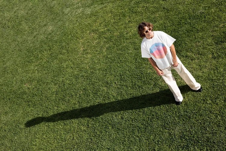 A young person standing on a grassy field, wearing sunglasses, a graphic T-shirt, and light-colored pants, casting a long shadow on the grass.