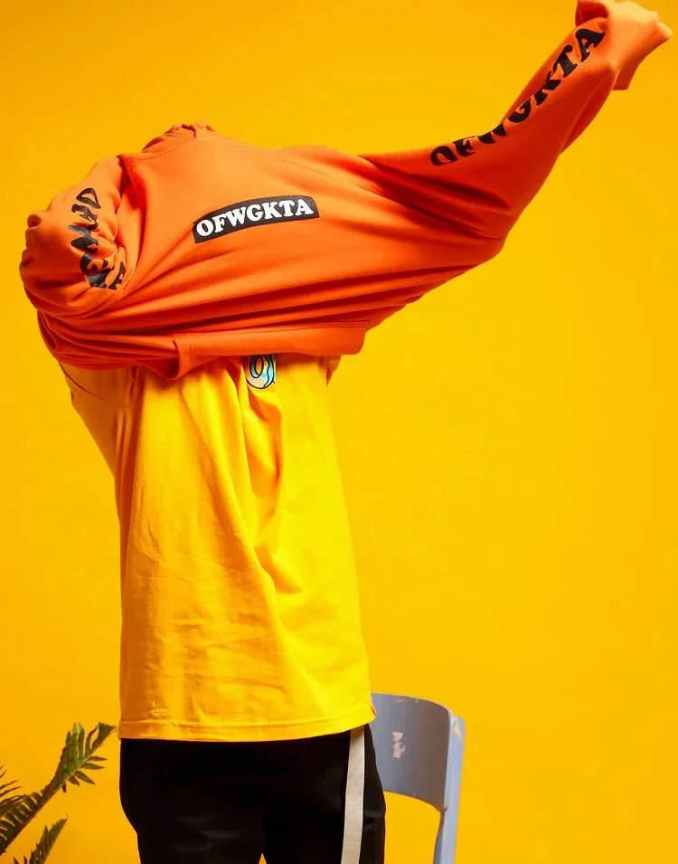 Person stretching in front of a yellow wall, wearing a yellow shirt and black pants, with orange sweatshirt covering their face and arm.