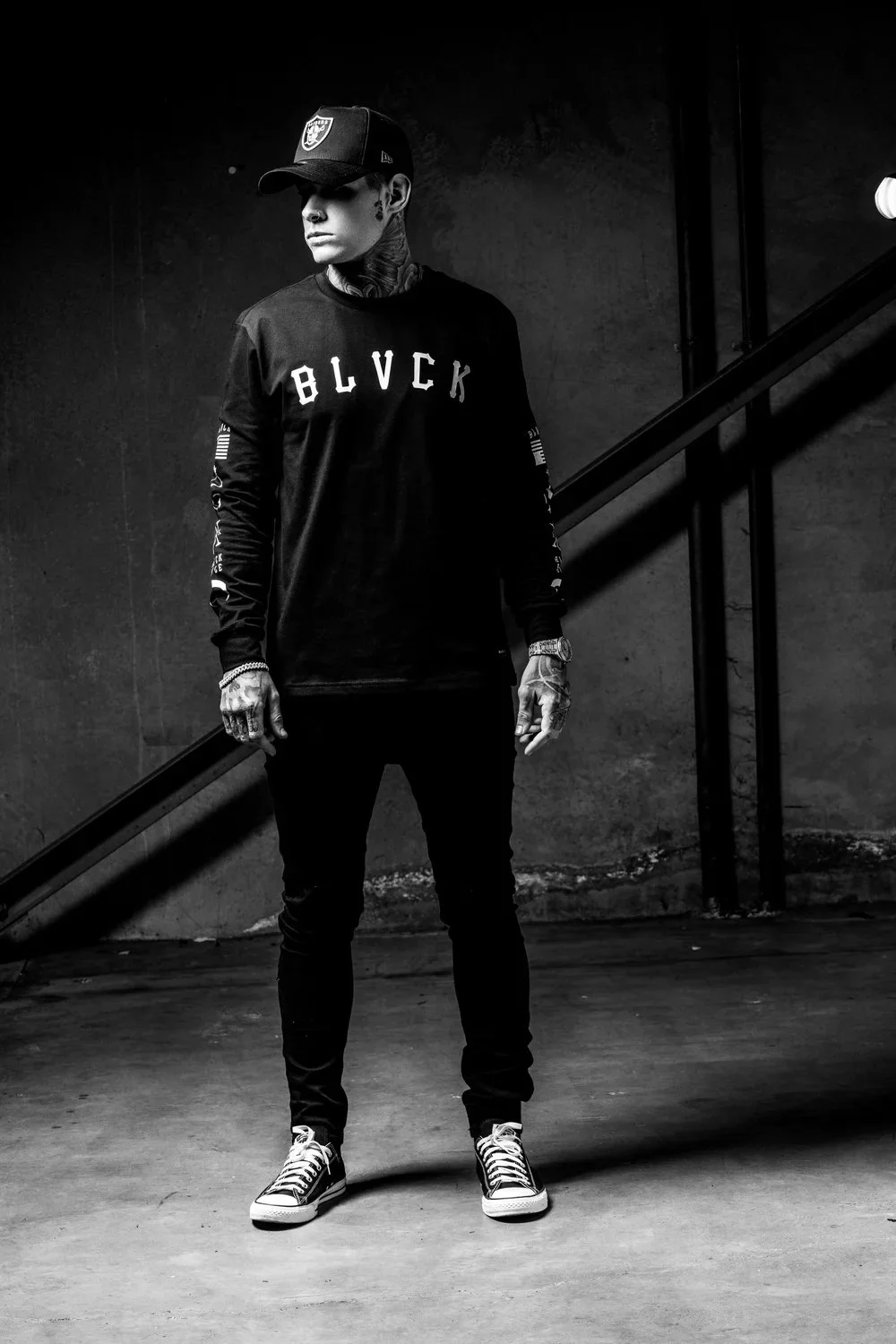 A young man with tattoos and tattoos on neck, wearing a black baseball cap, black long-sleeve shirt with 'BLACK' written on it, black pants, and black and white sneakers, standing against a dark textured wall in a studio setting.
