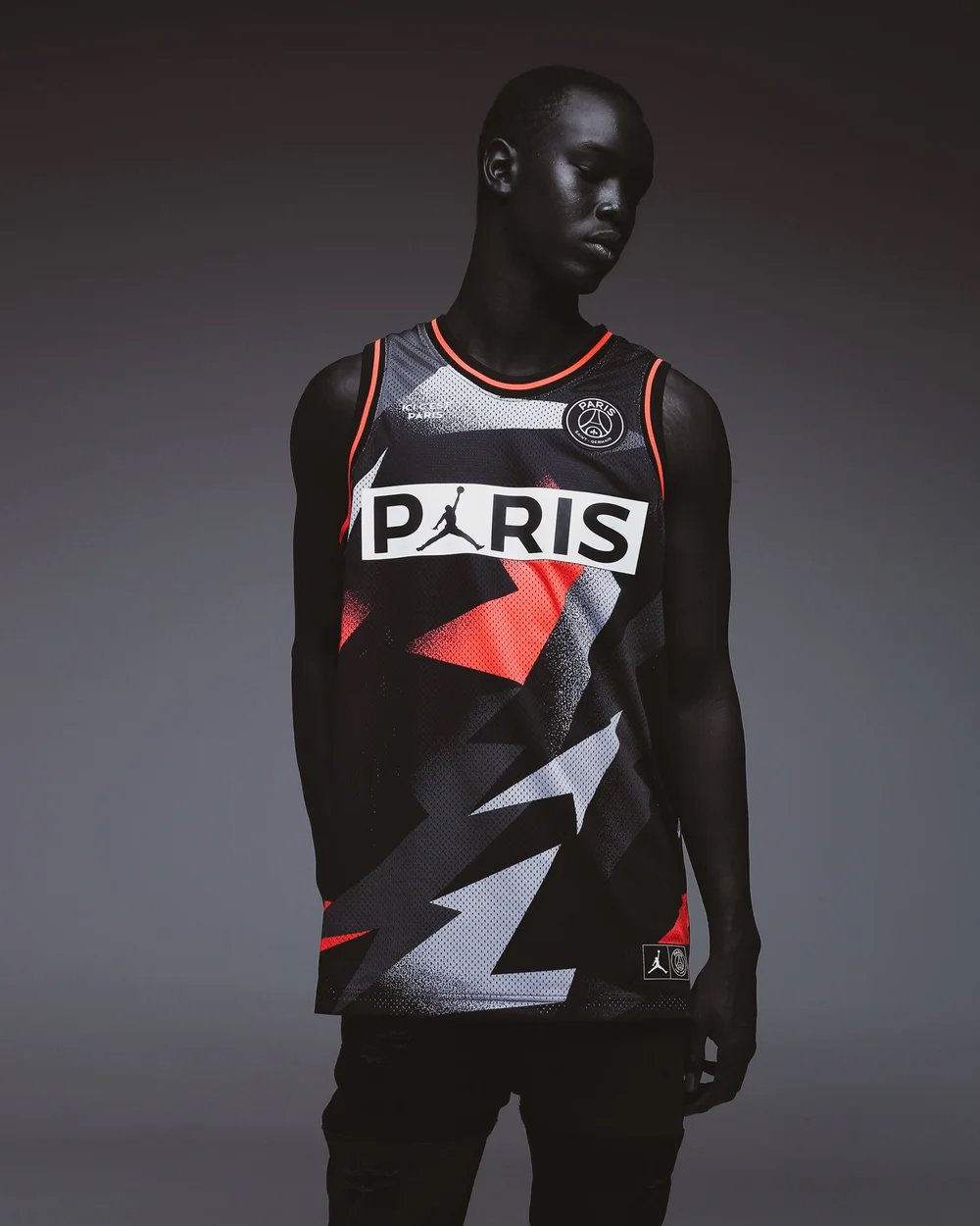 A woman with dark skin and short hair wearing a Paris Saint-Germain basketball jersey with black, red, and white geometric patterns.
