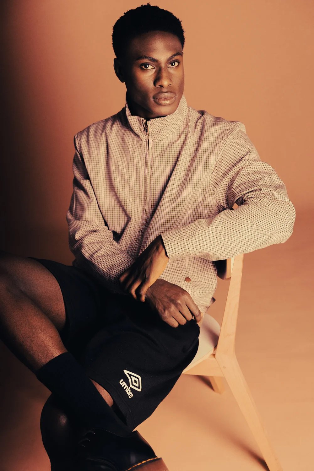 A young man with dark skin and short curly hair sitting on a wooden chair against a plain background. He is wearing a beige checkered zip-up jacket, black shorts, black socks, and black shoes. His expression is neutral, and he is looking directly at 