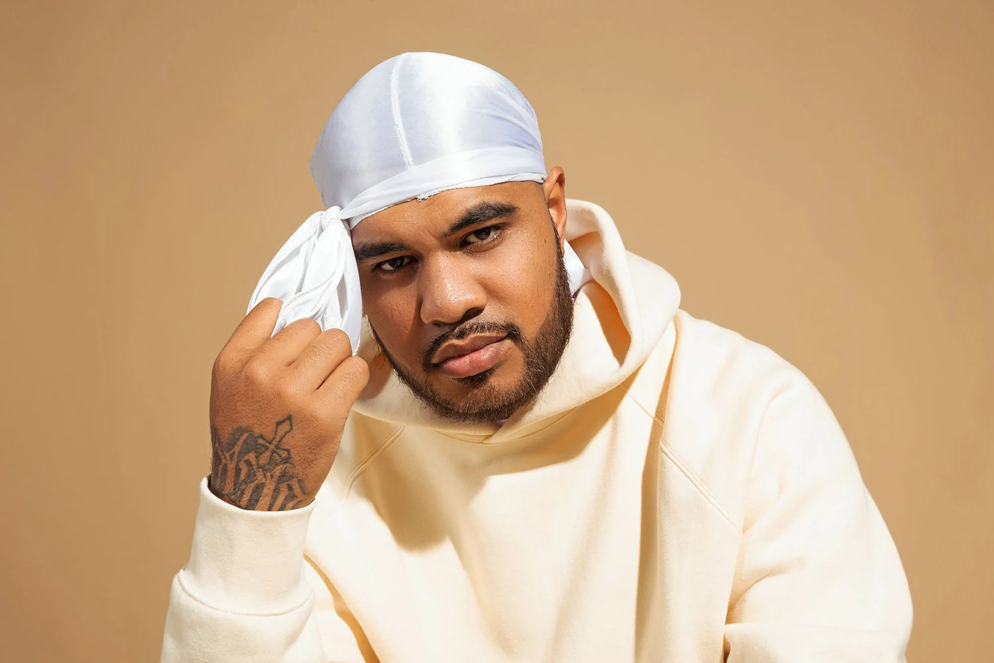 A man with a beard and tattoo on his hand, wearing a white hoodie and a white satin durag, is adjusting it with his right hand. He has a neutral expression and is posed against a plain beige background.
