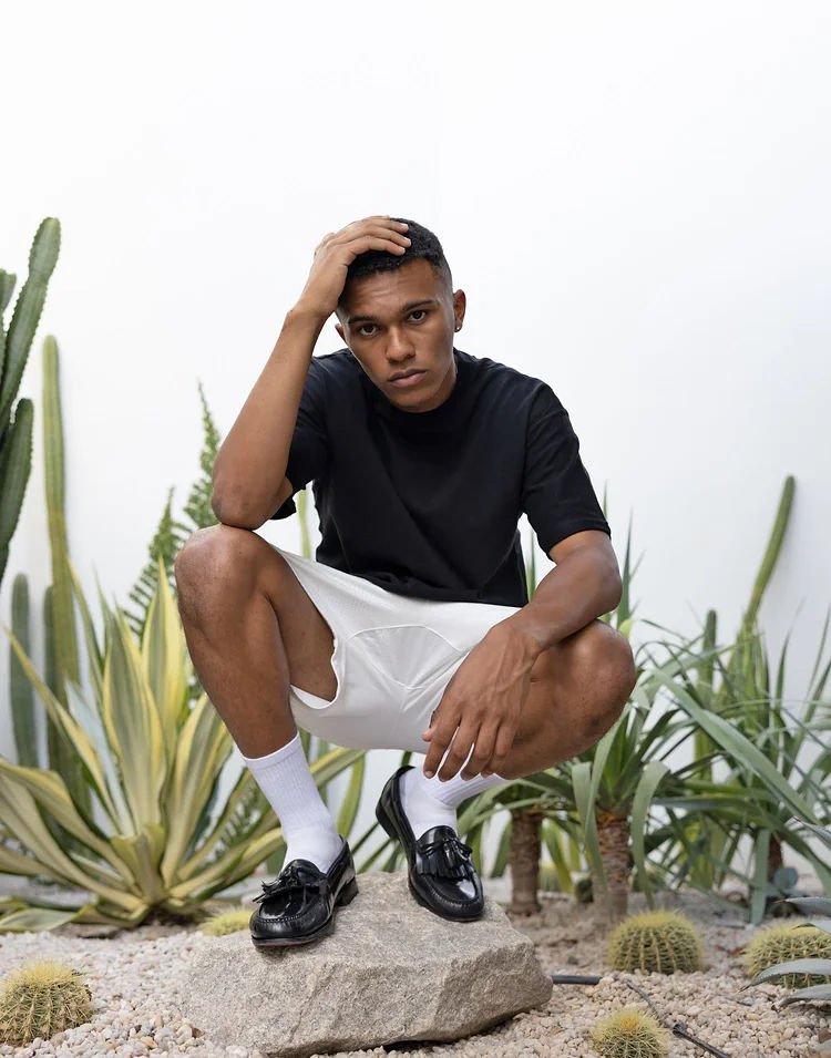 Young man crouching on a rock, touching his head, surrounded by desert plants and cacti, wearing a black T-shirt, white shorts, white socks, and black loafers.