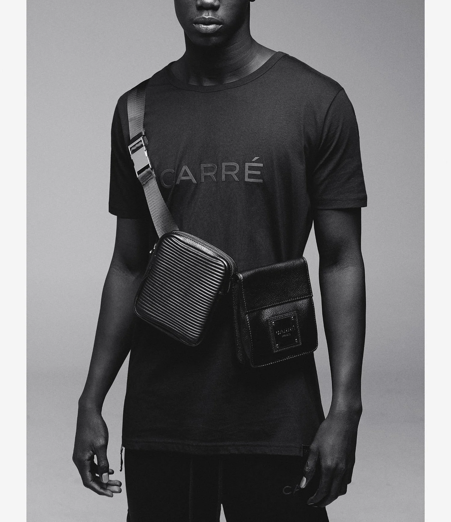 A person wearing a black T-shirt with the word 'CARRÉ' on it, carrying two black crossbody bags, one textured and one smooth, over a gray background.