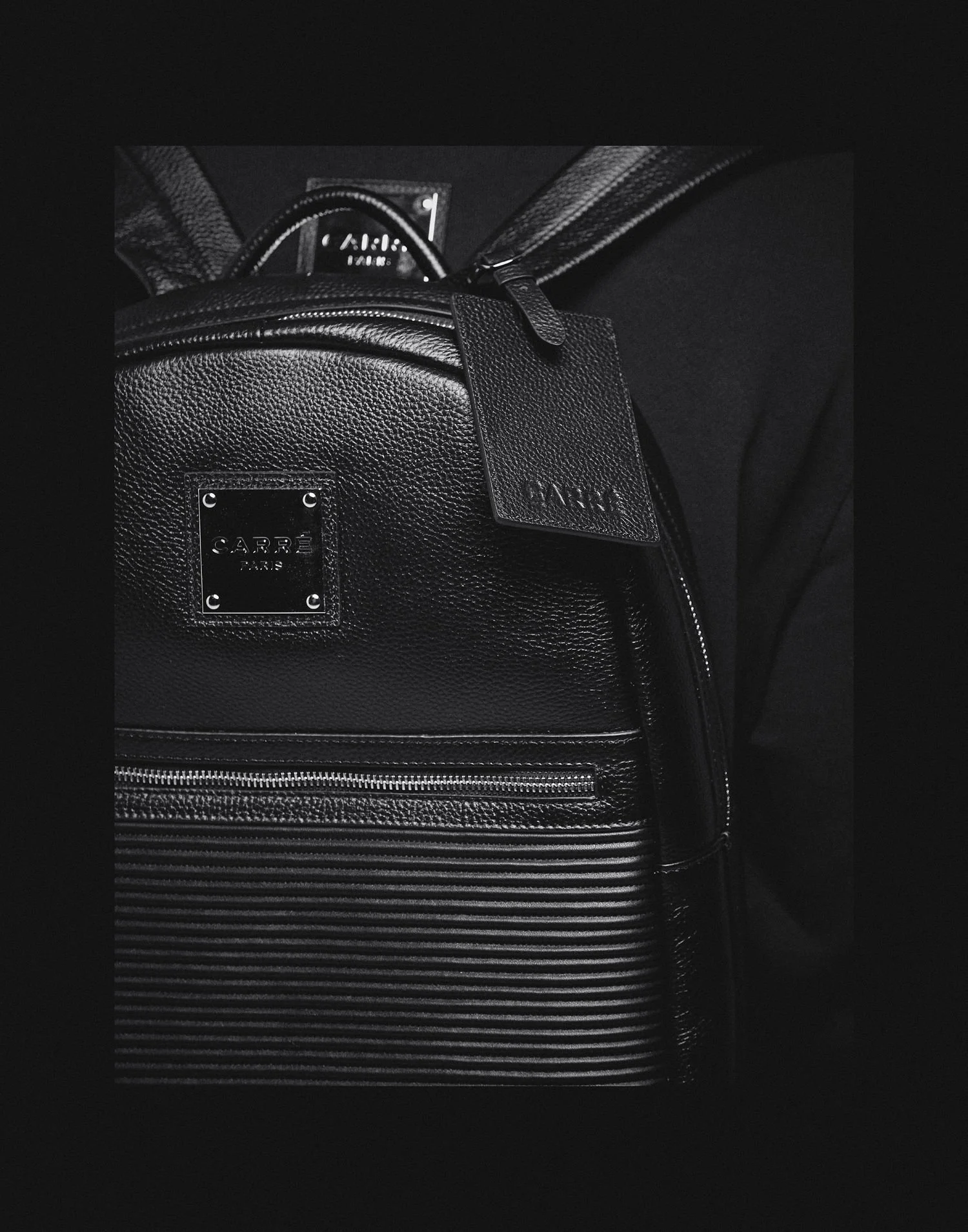Black Carré Paris backpack with leather texture, metal brand plate, and horizontal striped pattern on lower section, with a leather tag attached.