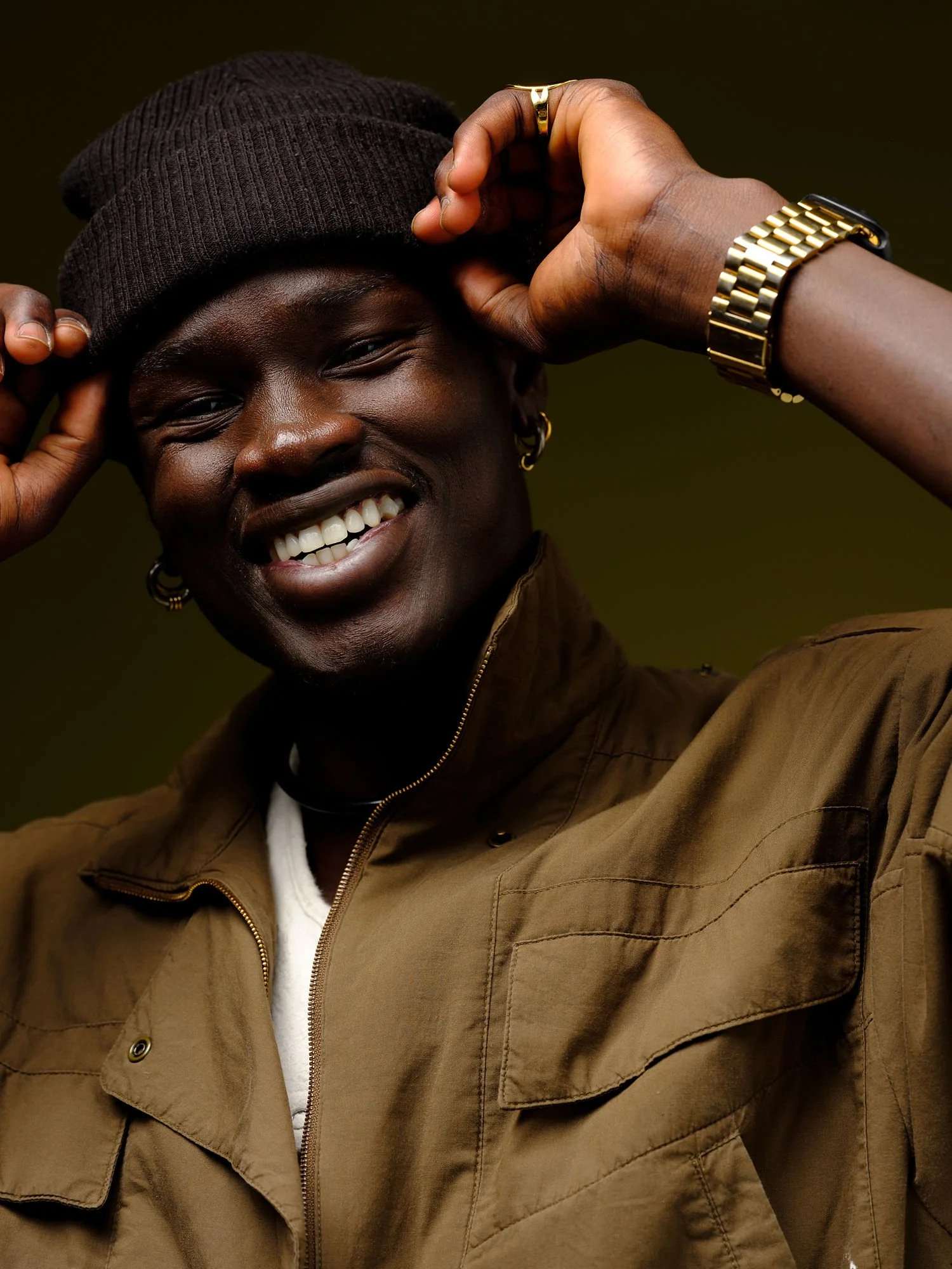 A smiling person with dark skin, wearing a brown beanie, gold jewelry, and a tan jacket, holding the beanie with both hands.