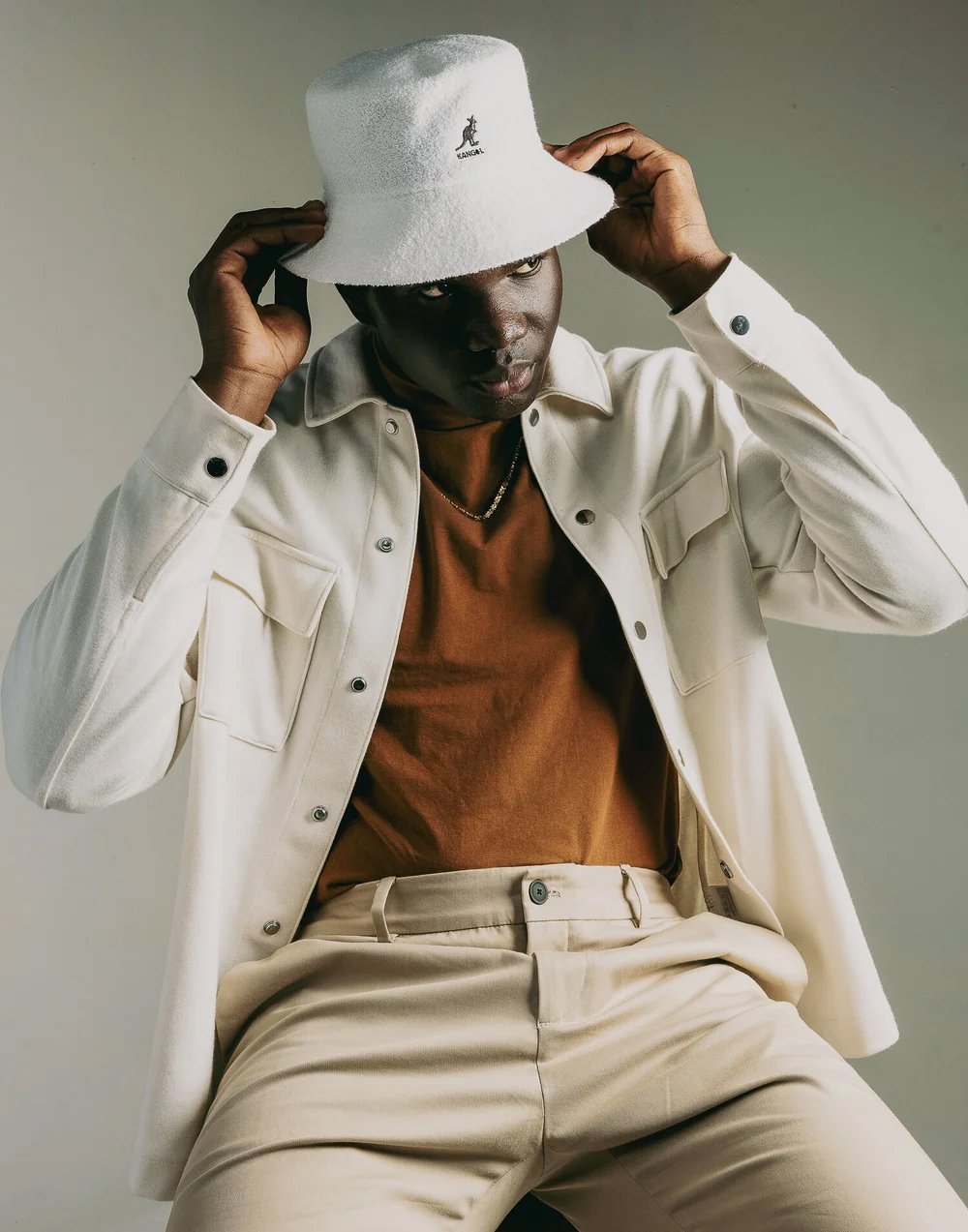 A man wearing a white bucket hat, a white jacket, a brown shirt, and beige pants, posing against a neutral background.