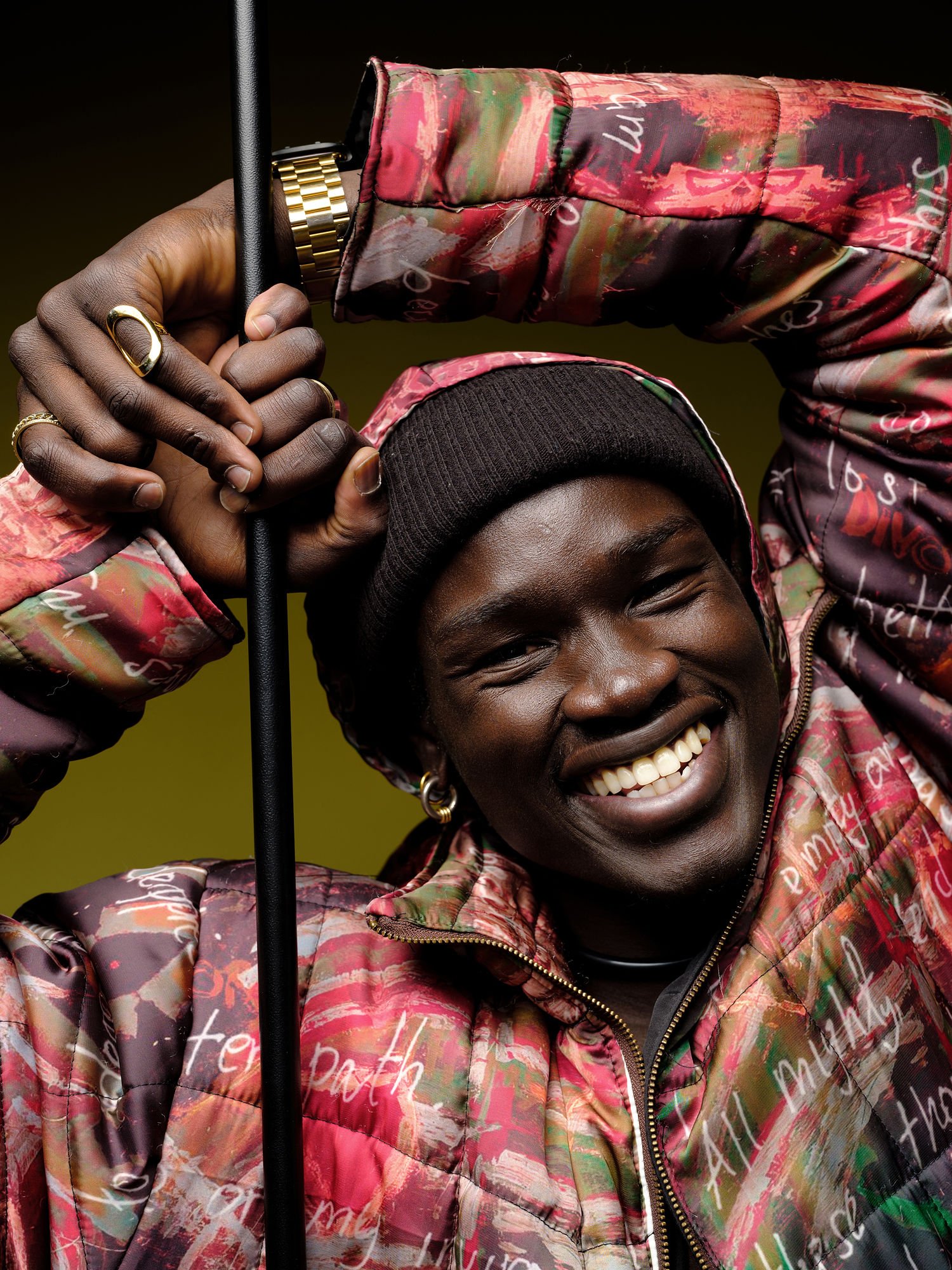 A smiling person wearing a colorful jacket, black beanie, and gold accessories, holding an umbrella or pole, against a yellow-green background.