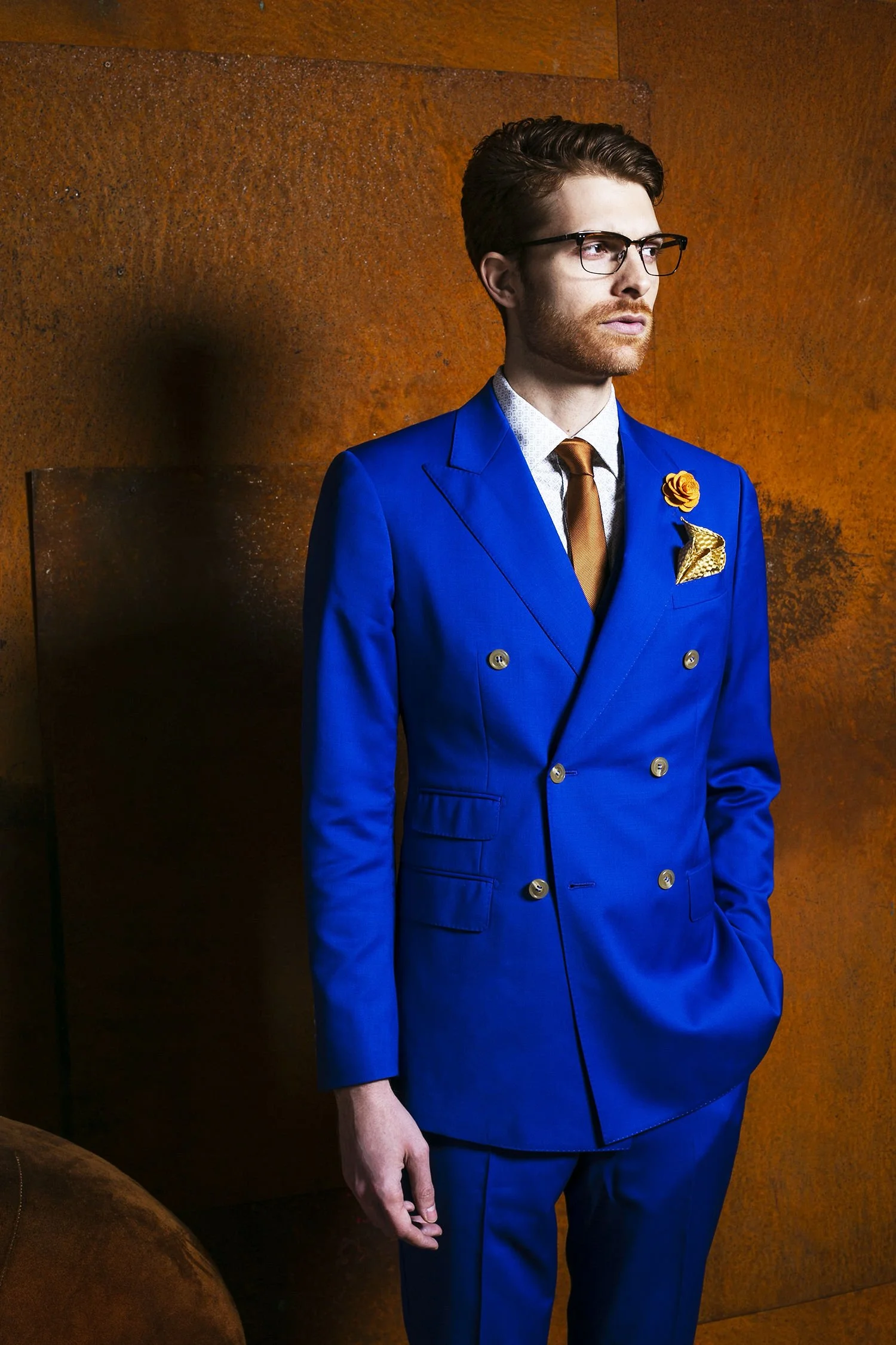 A man in a royal blue double-breasted suit, white shirt, bronze tie, and glasses stands against a rust-colored wall, with a yellow floral lapel pin and pocket square.