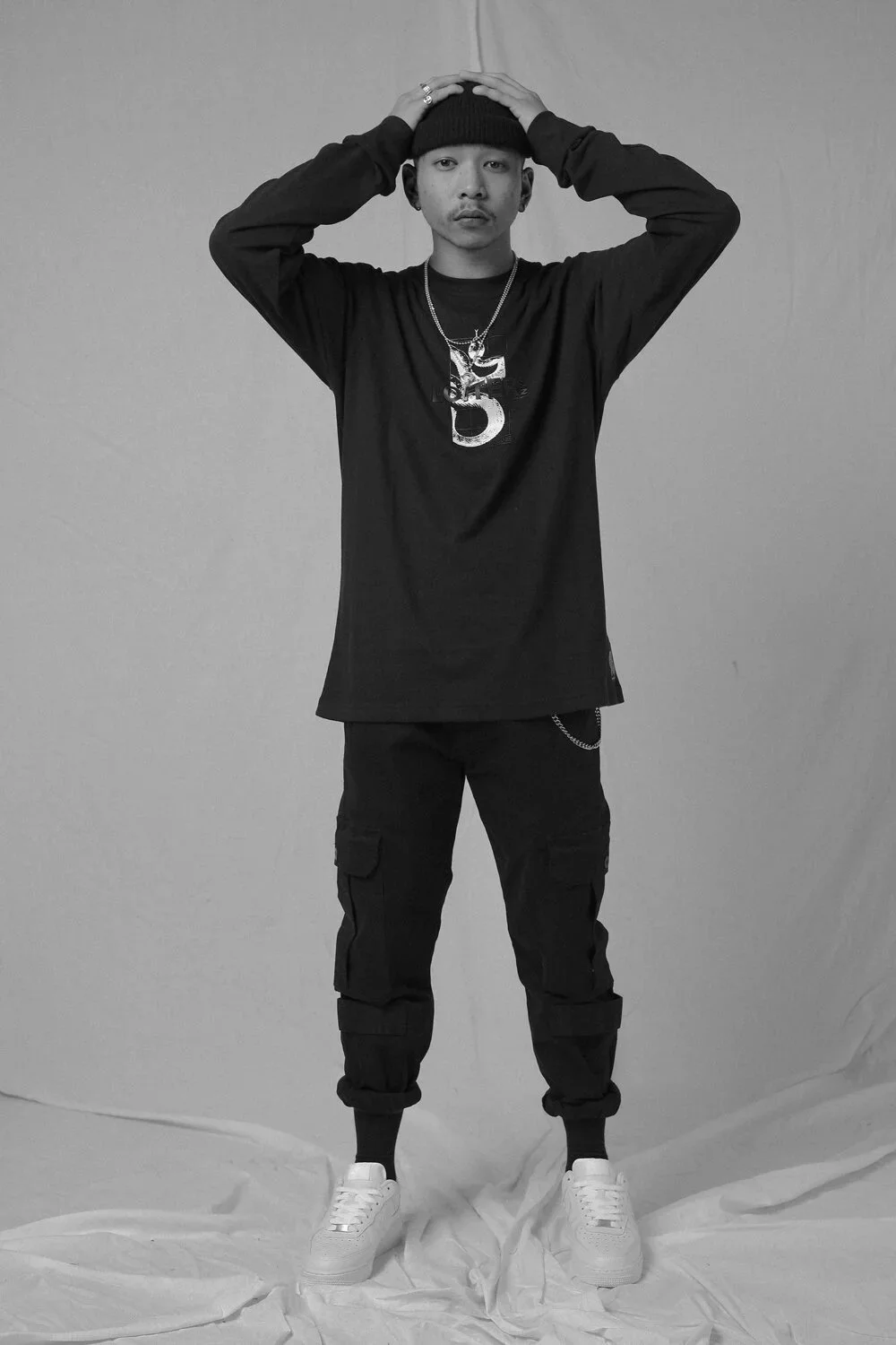 Black and white photo of a young man standing with his hands on his head, wearing a beanie, a long-sleeved shirt, cargo pants, and sneakers, against a plain backdrop.