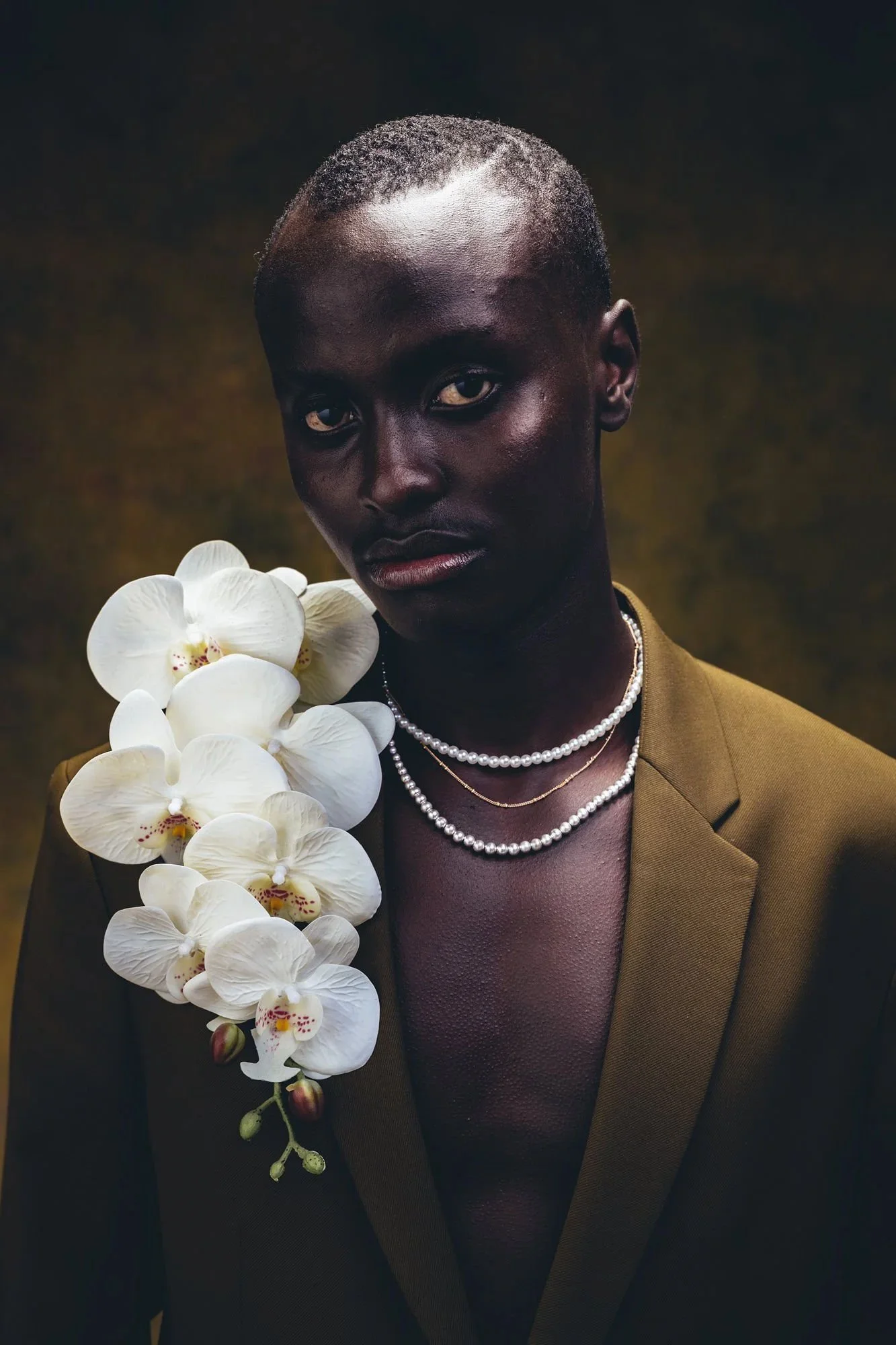Portrait of a person with dark skin, short hair, wearing a brown blazer, multiple pearl necklaces, and surrounded by white orchids.