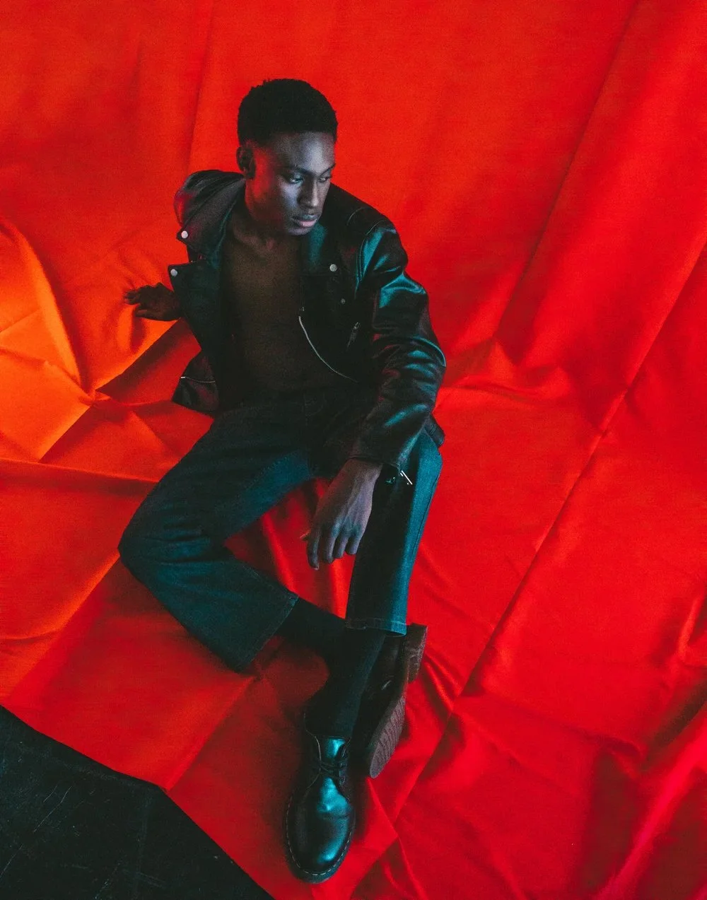 A young man with short hair, wearing a black leather jacket, black pants, and black boots, sitting against a vibrant red fabric backdrop, looking off to the side with a serious expression.