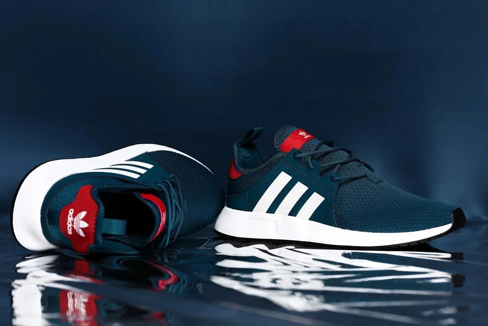 Pair of Adidas sneakers, one lying down and the other standing, on a reflective surface with a dark background.