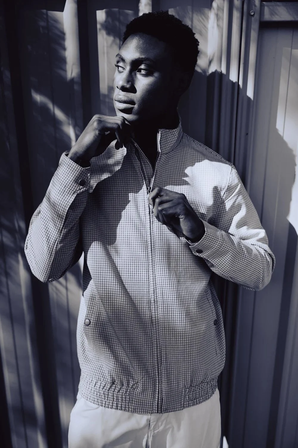 Young man in checkered zip-up jacket standing outdoors against a corrugated metal background, shadowed by sunlight.