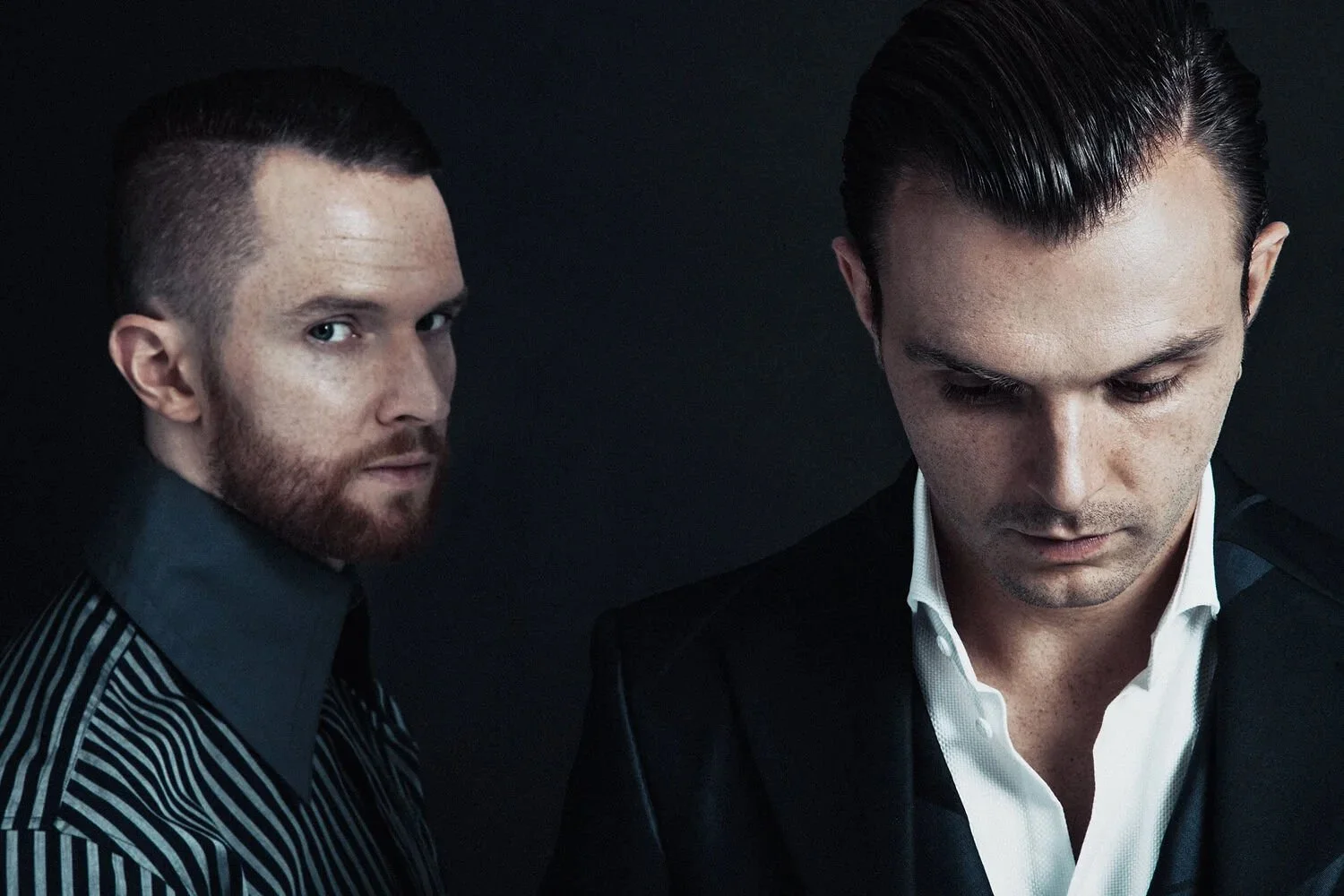 Two men with slicked-back hair and serious expressions, dressed in dark clothing, against a black background.