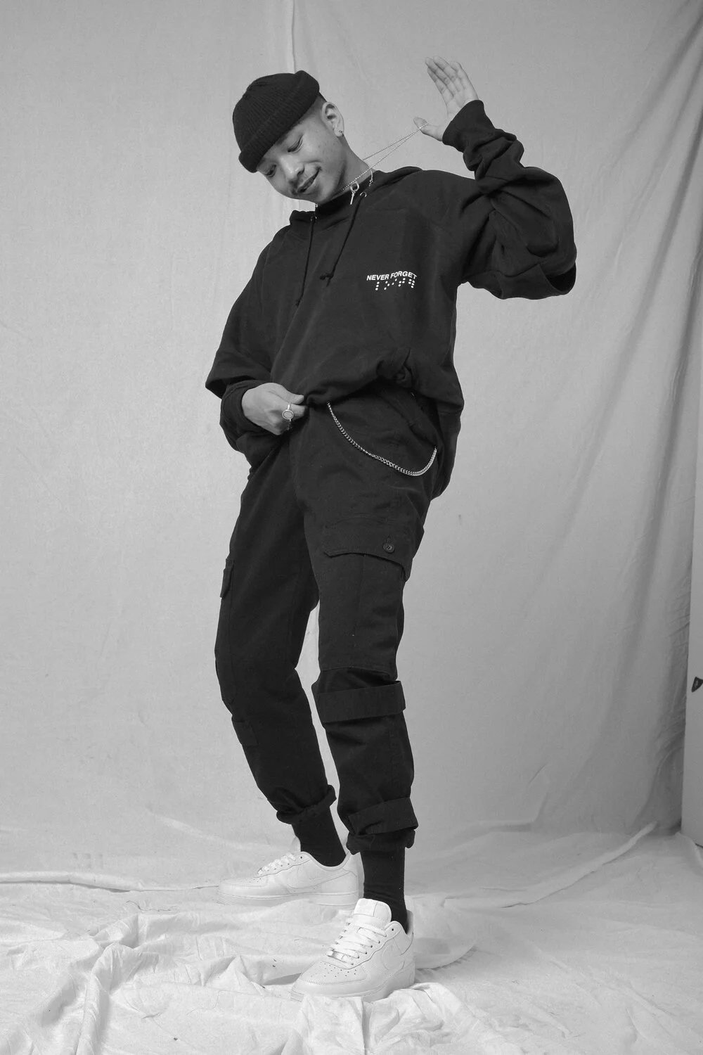 A young man in casual streetwear, including a beanie, hoodie, cargo pants, and sneakers, standing in front of a plain backdrop. He is posing with a relaxed demeanor, holding a chain with one hand and touching his hoodie with the other. The image is i