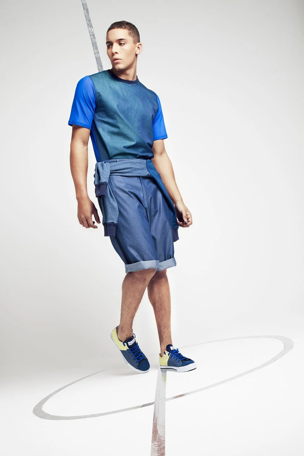 A young man wearing a blue T-shirt, denim shorts, and sneakers standing inside a basketball court with a white background.
