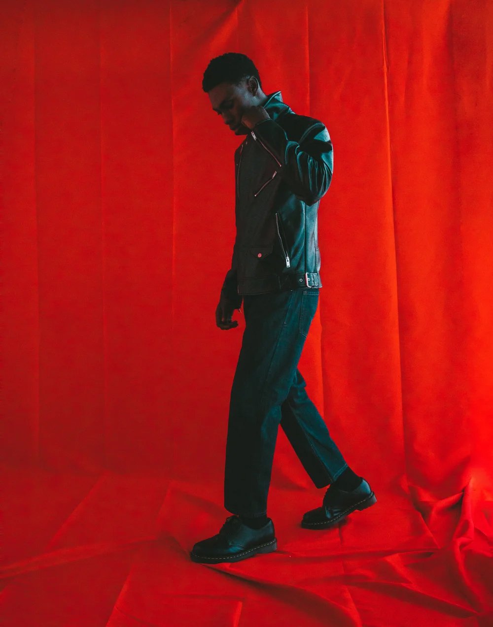 A man dressed in black leather jacket, black jeans, and black boots standing against a red curtain backdrop, with his head slightly bowed and one hand touching his neck.