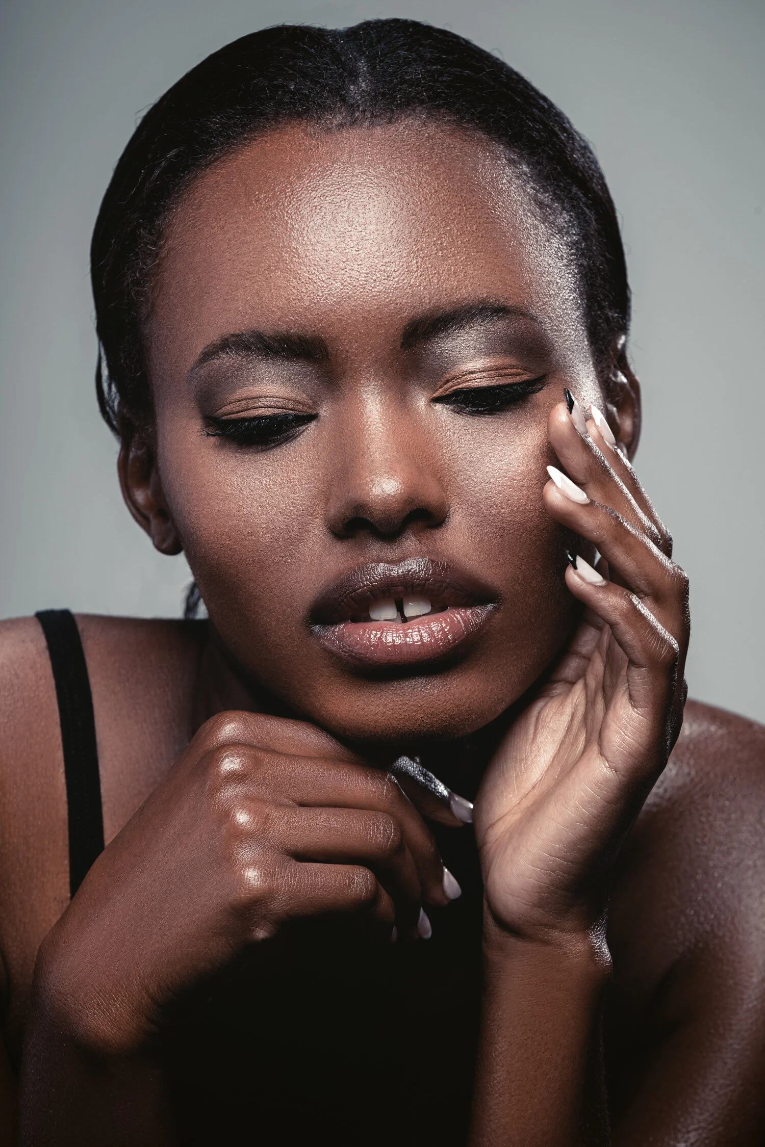 Close-up portrait of a woman with dark skin, smooth skin, dark eye makeup, and glossy lips, touching her face with her hand.