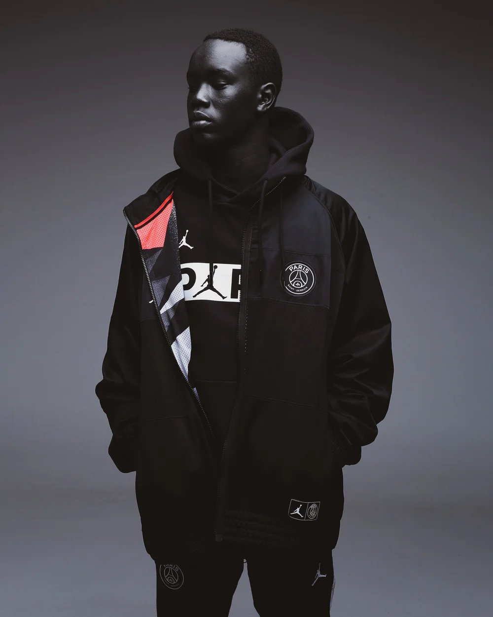 A young man with dark skin and short hair wearing a black jacket, hoodie, and pants with Paris Saint-Germain logos, standing against a gray background.