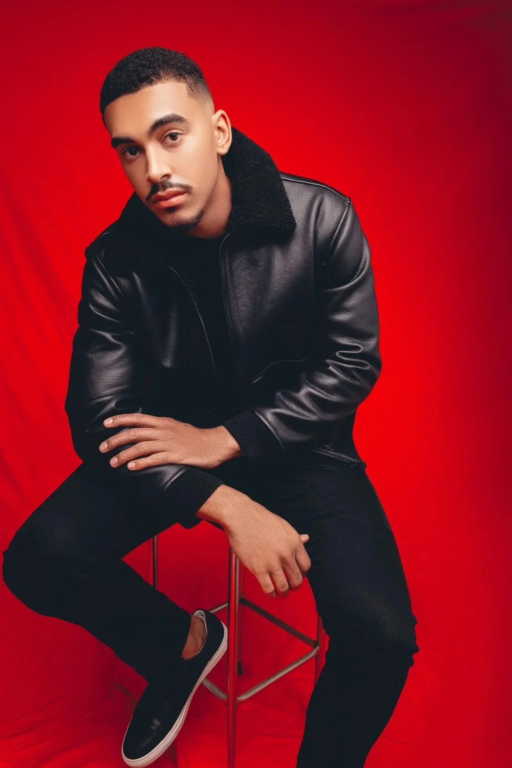 Young man with short hair and mustache sitting on a stool against a red backdrop, wearing a black leather jacket and black pants.