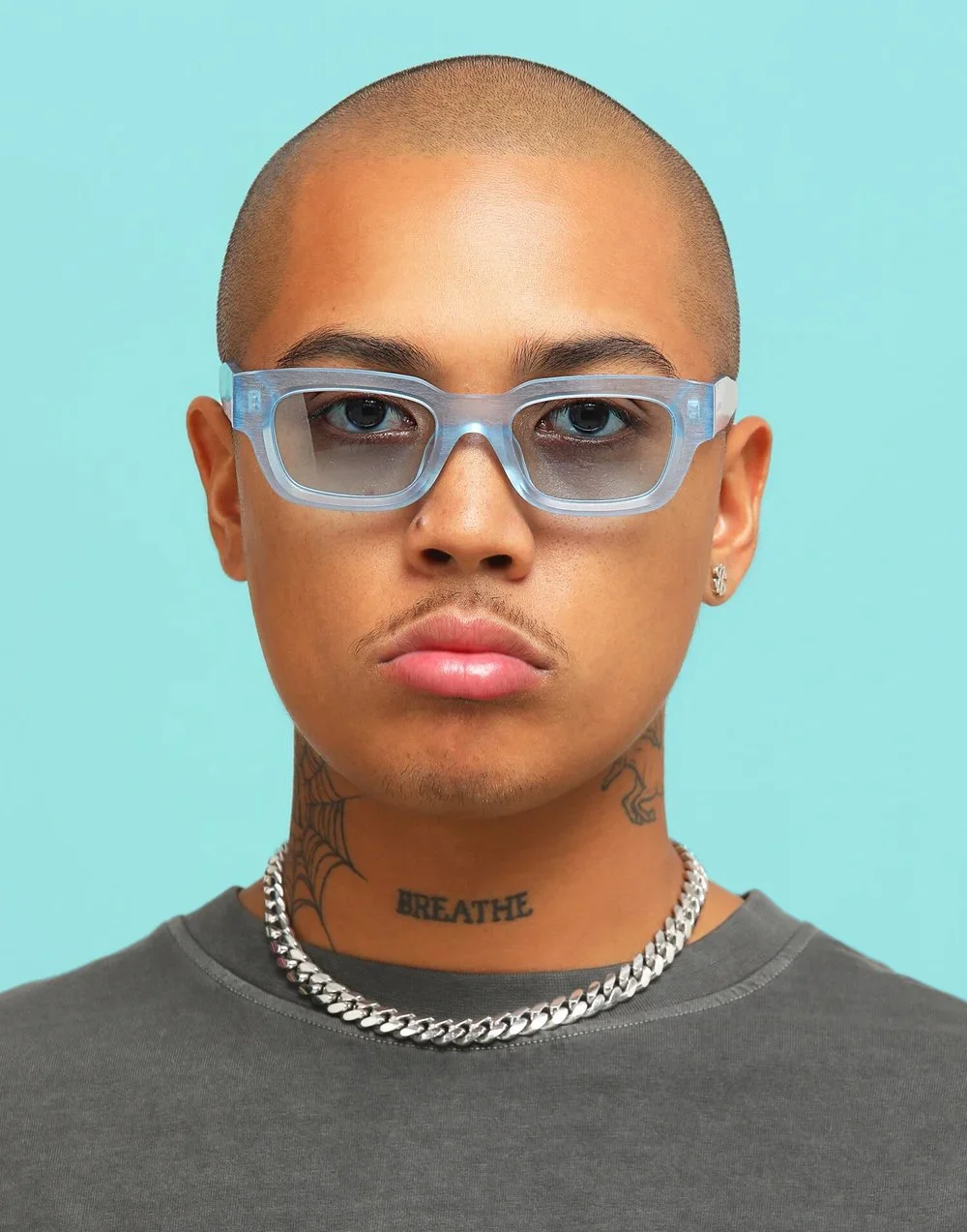 A person with a buzz cut, wearing clear glasses and a silver chain necklace, has tattoos including the word 'BREATHE' on the neck and a spider web tattoo near the shoulder, against a light blue background.